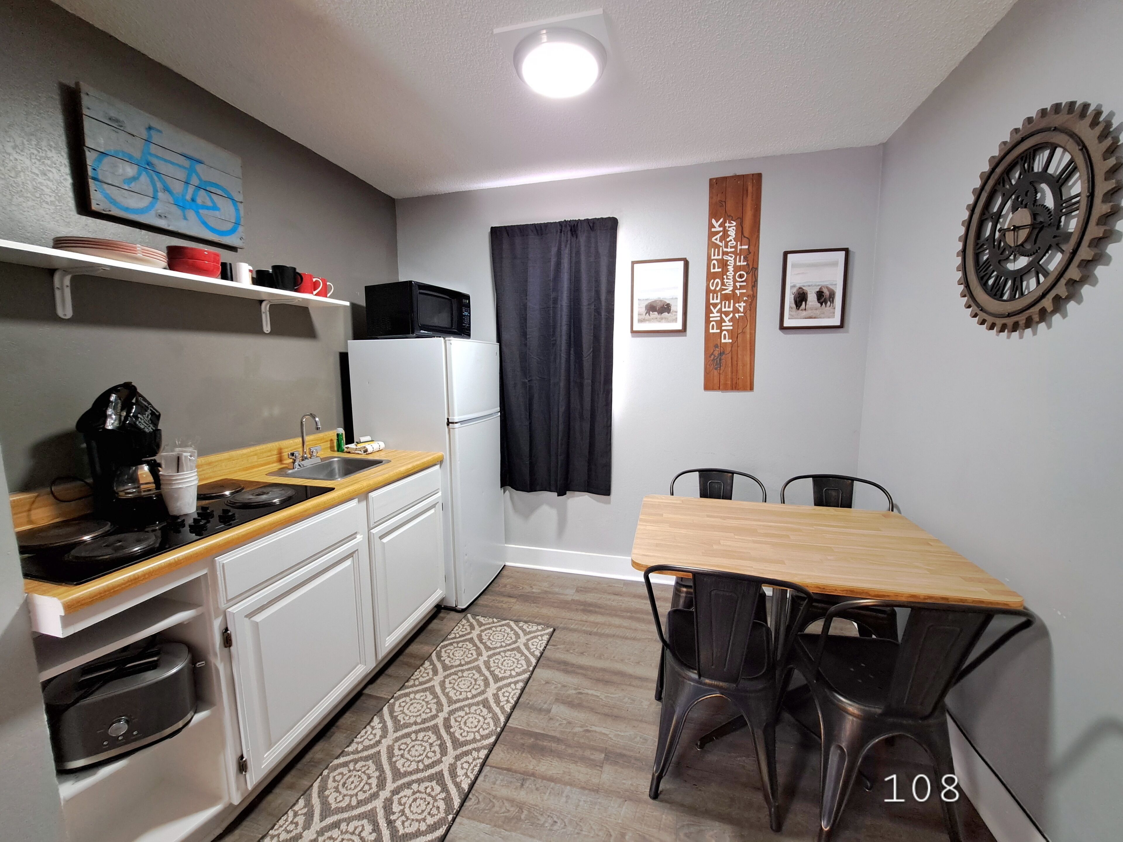 Een kleine keuken met witte kastjes, een magnetron en een koffiezetapparaat. Er is een eettafel met stoelen en een vloerkleed.