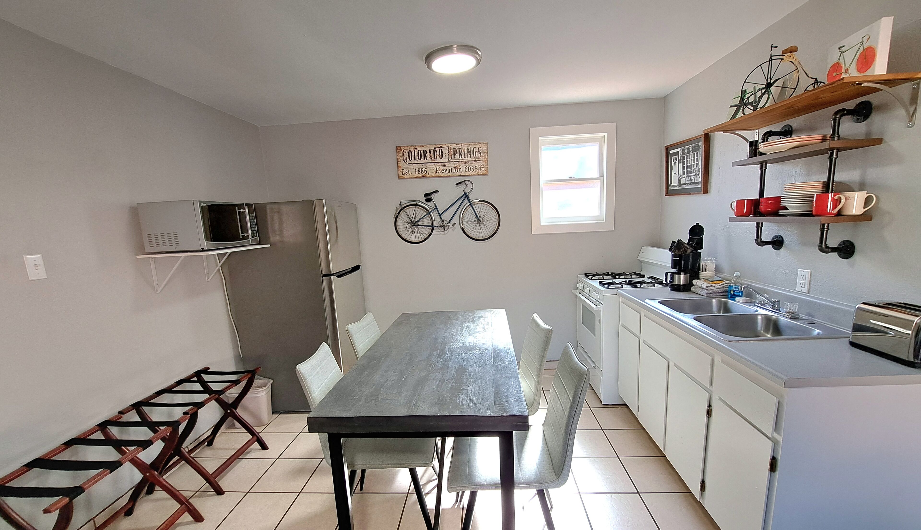 Una cocina funcional con mesa y sillas, microondas, refrigerador, estufa y lavamanos. Se observan estantes con tazas y una decoración de bicicleta en la pared.