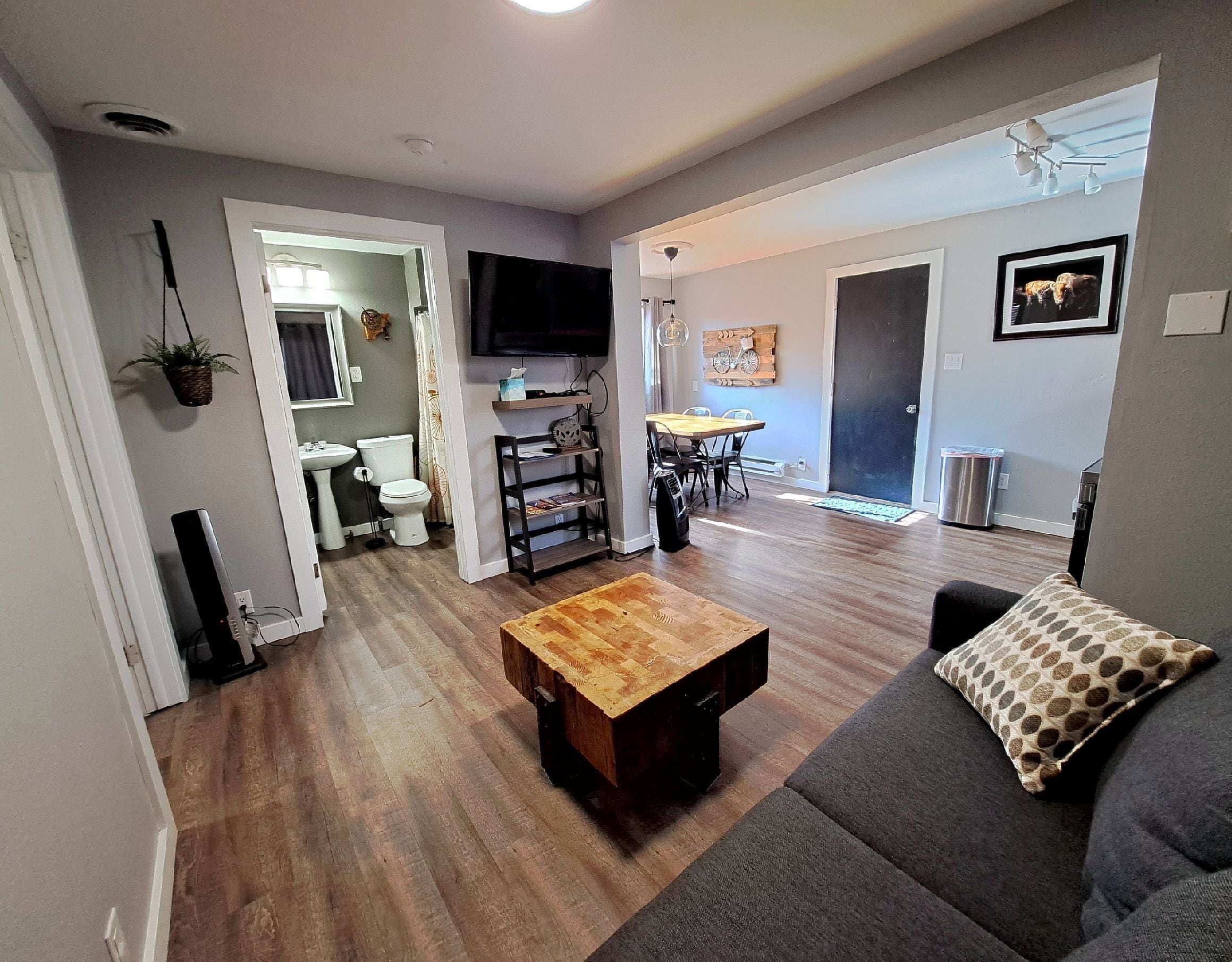 Un salon avec une table basse en bois, un canapé gris agrémenté d’un coussin à motifs et un téléviseur fixé au mur.