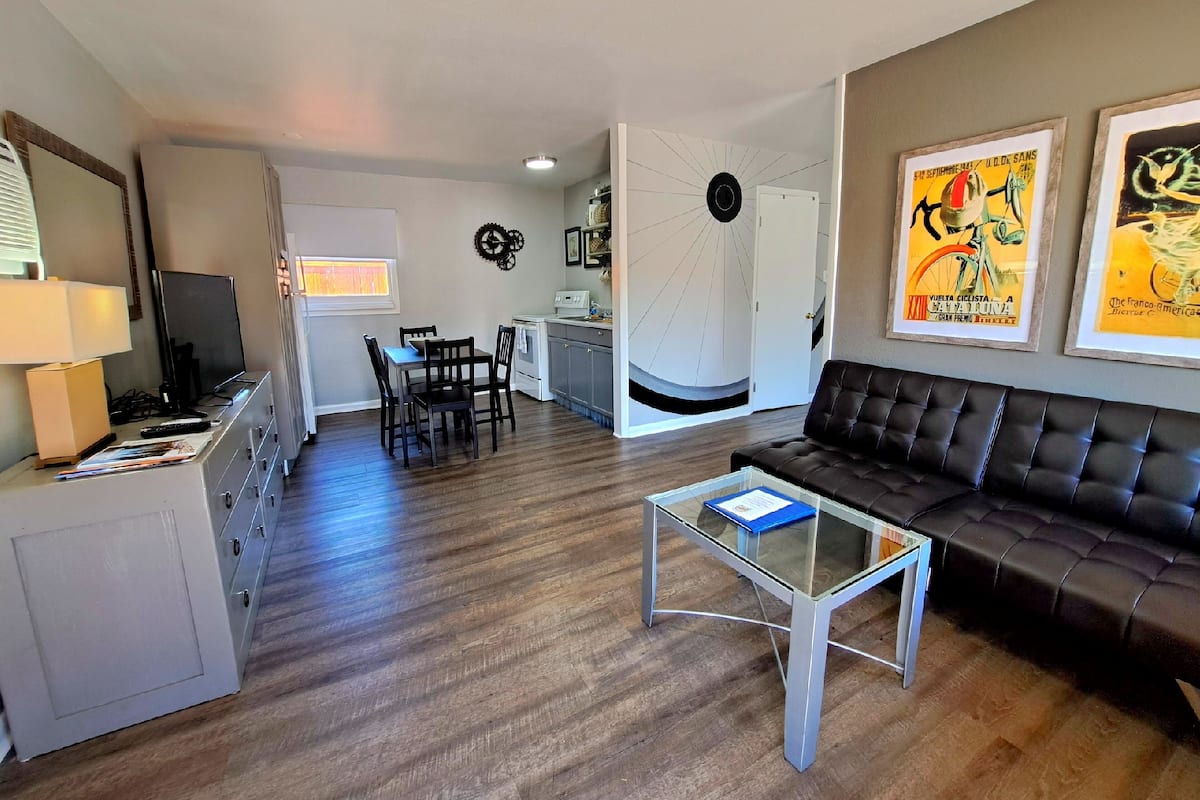 A modern living room with a leather sofa, a glass coffee table, and a dining area with a table and chairs. There is a kitchen area with a stove and cabinets.