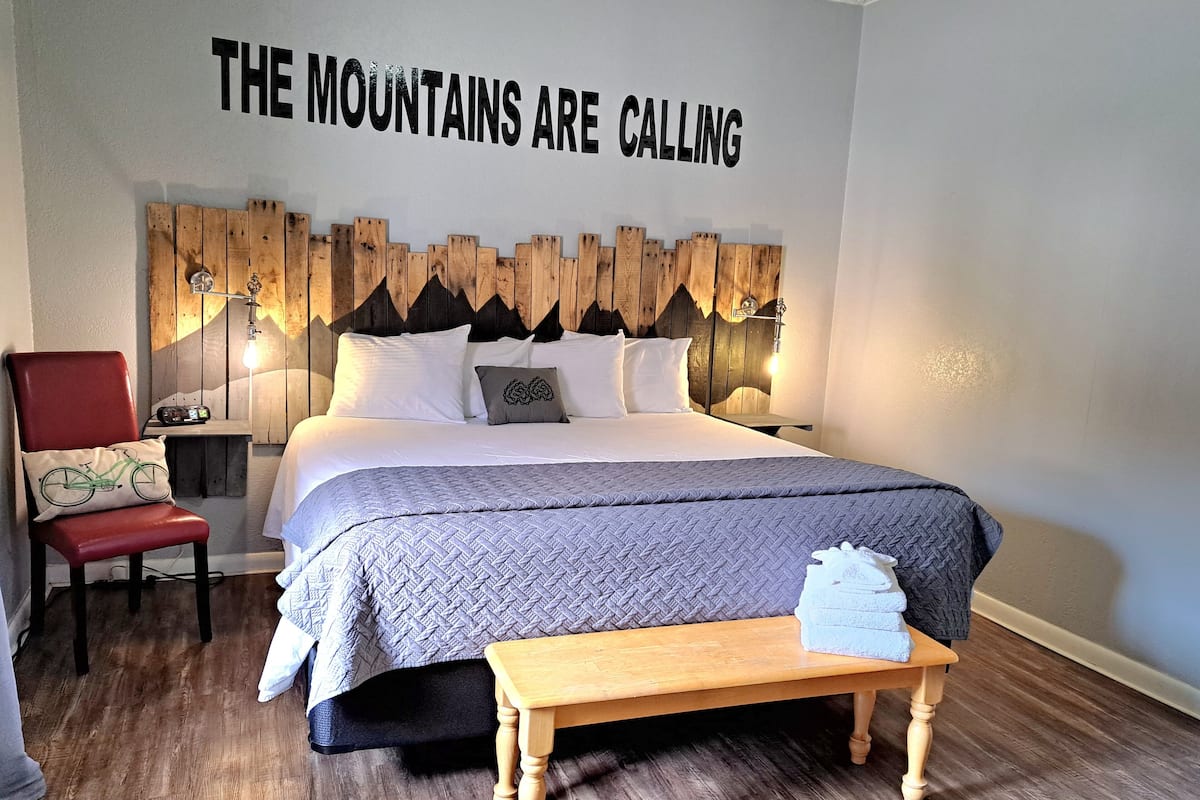 A bedroom with a bed, a chair, a wooden headboard, and a wall sign that reads "THE MOUNTAINS ARE CALLING".