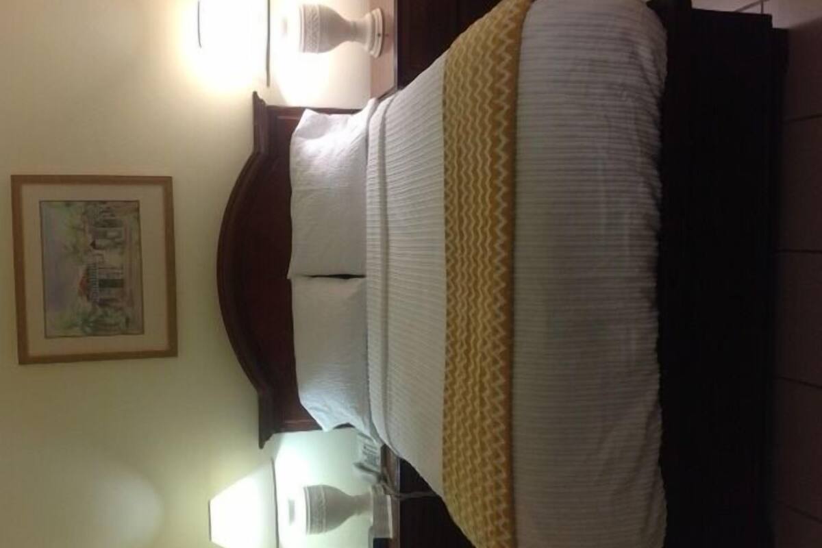 A neatly made bed with a white and yellow striped blanket, a framed picture on the wall, and a bedside lamp.