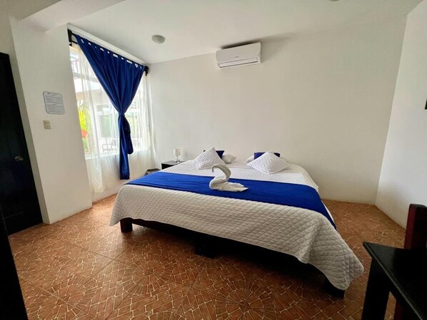 A hotel room with a bed, a blue curtain, a white air conditioning unit, and a tiled floor.