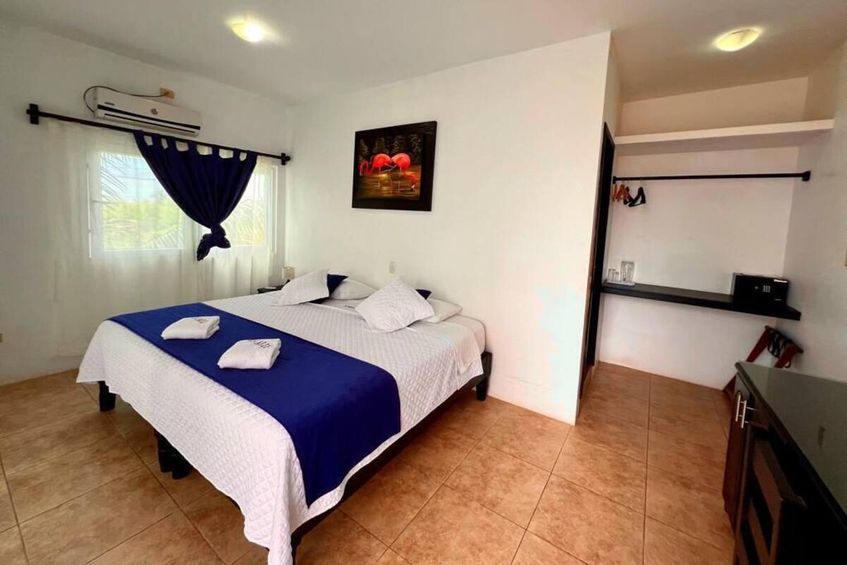A hotel room with a bed, a window with curtains, an air conditioning unit, a painting on the wall, a small shelf with items, and a desk with a chair.