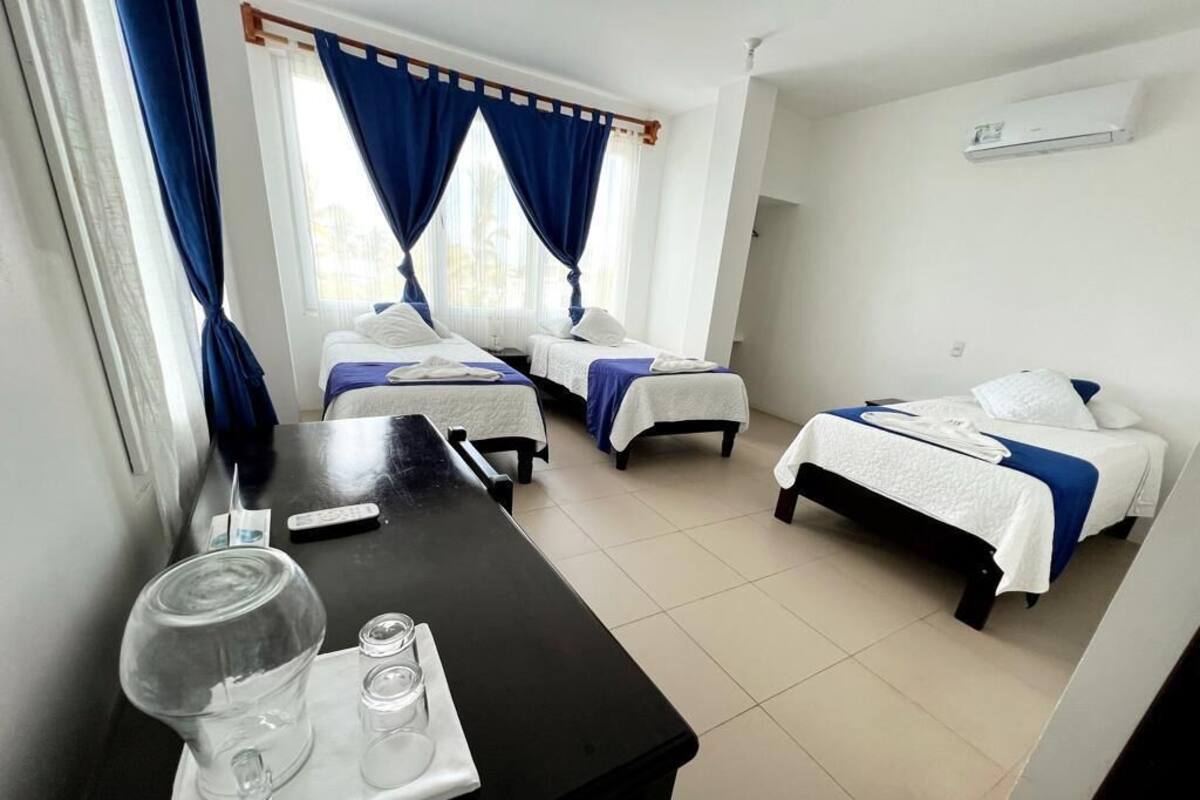 A hotel room with two single beds, a small table with a glass pitcher and glasses, and a window with blue curtains.