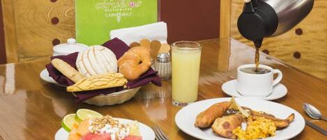 A breakfast table with croissants, a basket of bread, a plate with watermelon and yogurt, a glass of juice, and a cup of coffee being poured.