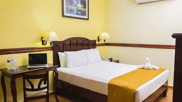 A hotel room with a bed, a desk with a laptop, two lamps, a framed picture on the wall, and an air conditioner.