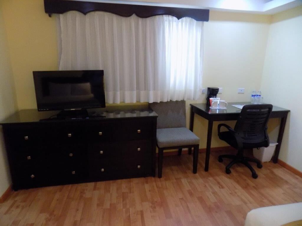 A small, furnished room with a television, a desk with a chair, a sofa, and a window with curtains.