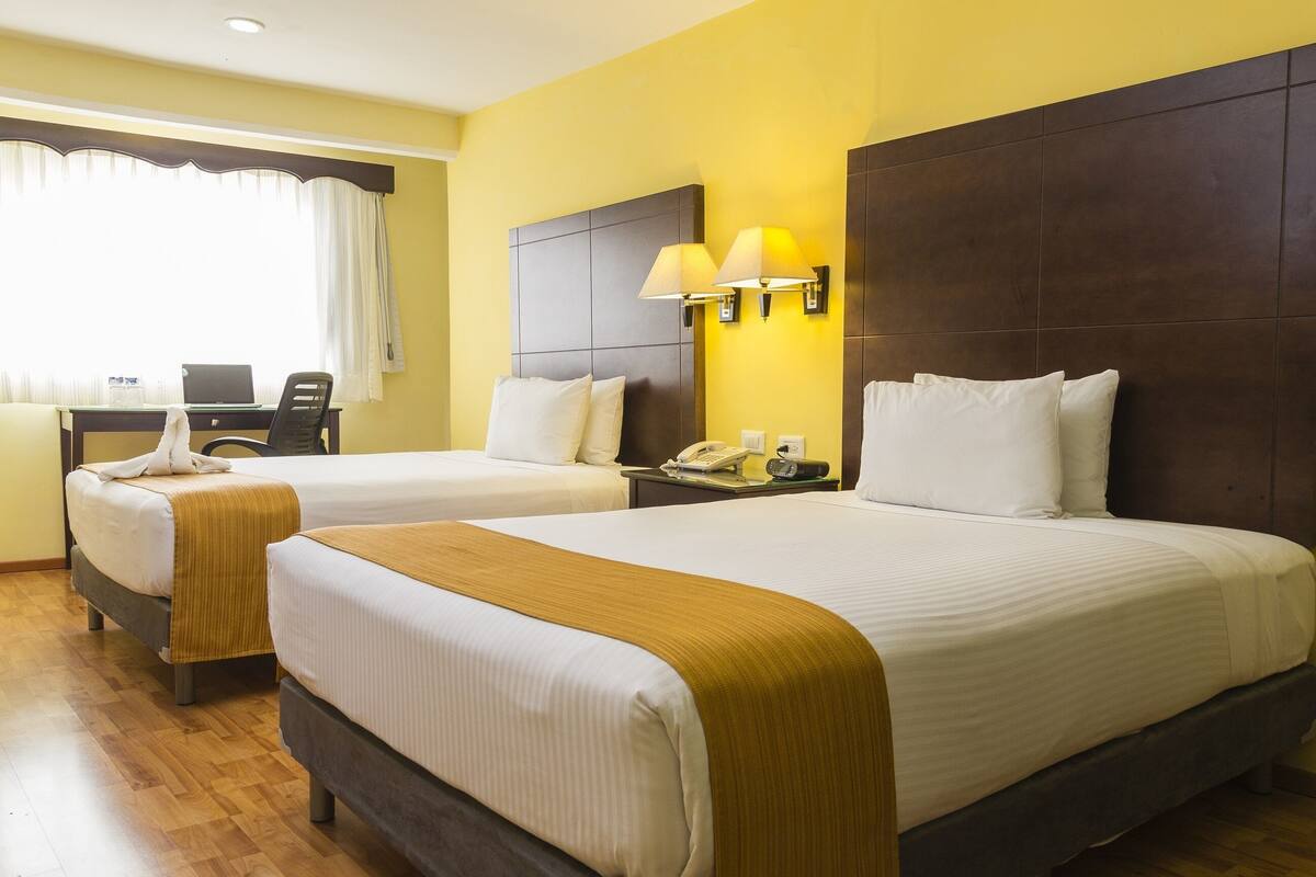 A hotel room with two beds, a desk with a laptop, a telephone, and a window with curtains.