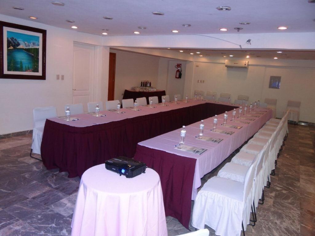 Un salón de conferencias con una larga mesa, sillas blancas y un proyector sobre una mesita.