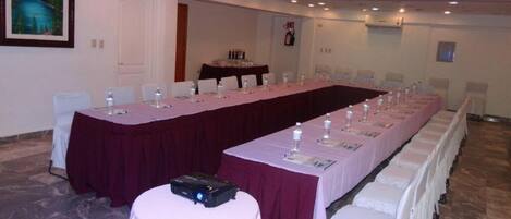 Un salón de conferencias con una larga mesa, sillas blancas y un proyector sobre una mesita.