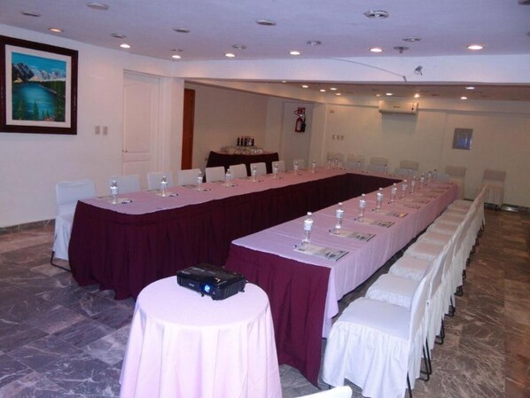 Une salle de conférence avec une longue table, des chaises blanches et un projecteur posé sur une petite table.