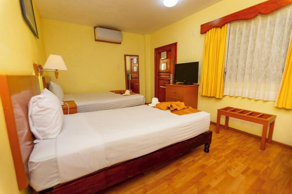 A hotel room with two single beds, a wooden nightstand, a television, and a window with yellow curtains.