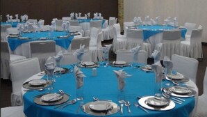 Un salón de banquetes con mesas redondas cubiertas con manteles azules, sillas blancas y cubiertos dispuestos para un evento formal.