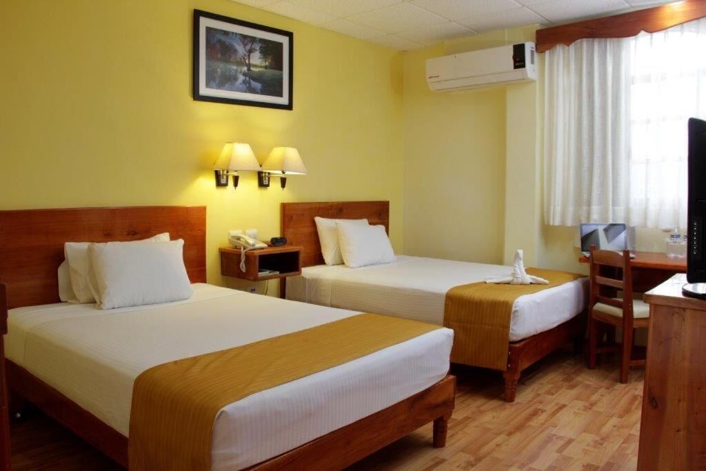 A hotel room with two beds, a desk, a chair, a TV, and an air conditioning unit.