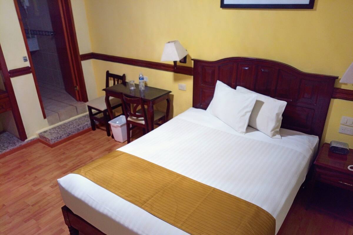 Habitación de hotel con cama, cabecera de madera, una mesa con sillas, lámpara y un cubo de basura.