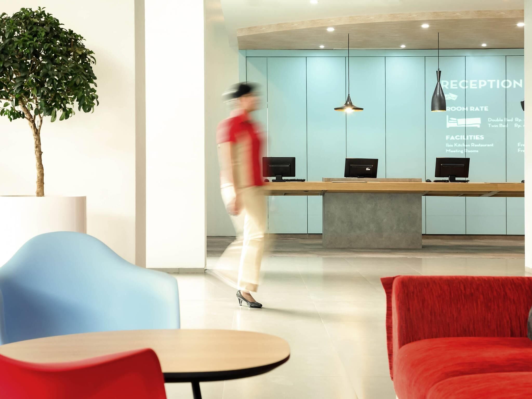 Un mostrador de recepción con pantalla digital, una planta y un área de descanso con sillas rojas y azules.