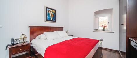 Een hotelkamer met een houten bed, witte beddengoed, een rode plaid, een nachtkastje met een lamp, een schilderij aan de muur en een raam met uitzicht.