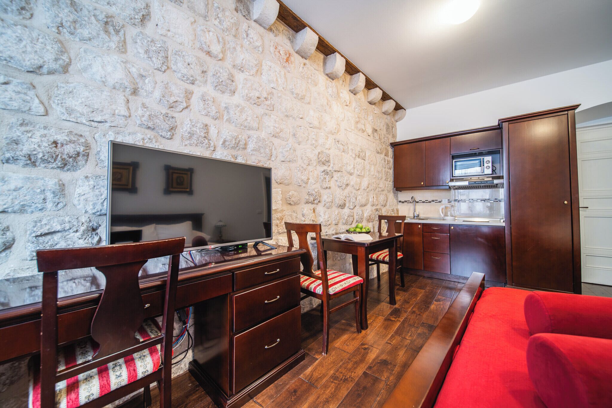 Habitación con pared de piedra, muebles de madera, televisor de pantalla plana, sofá rojo y una zona de cocina.