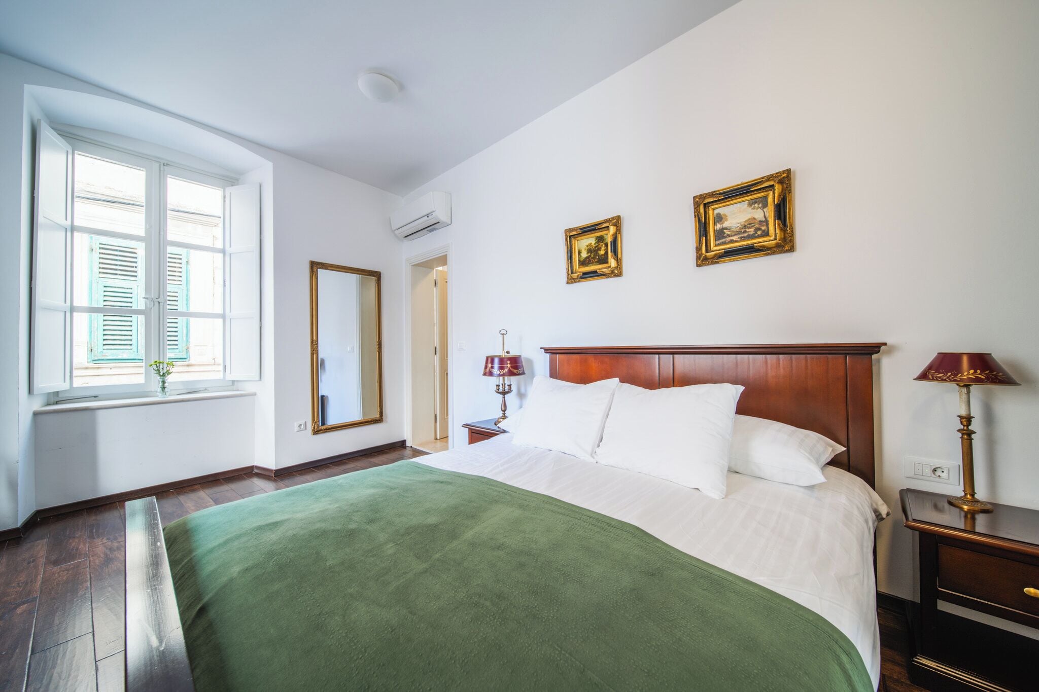 A hotel room with a large bed, a wooden headboard, a nightstand with a lamp, a window with shutters, and two framed pictures on the wall.