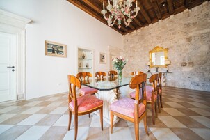 A dining area with a glass table surrounded by wooden chairs with patterned cushions, a chandelier, a stone wall, and a mirror on the wall.