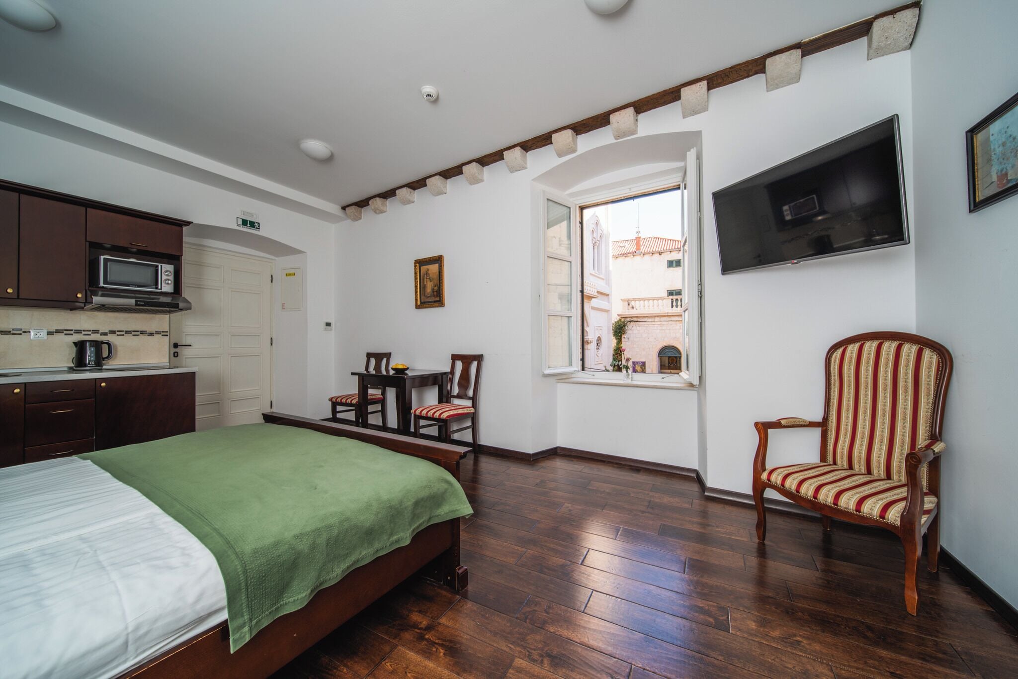 A room with a bed, a chair, a table, a TV, and a window with a view of buildings outside.