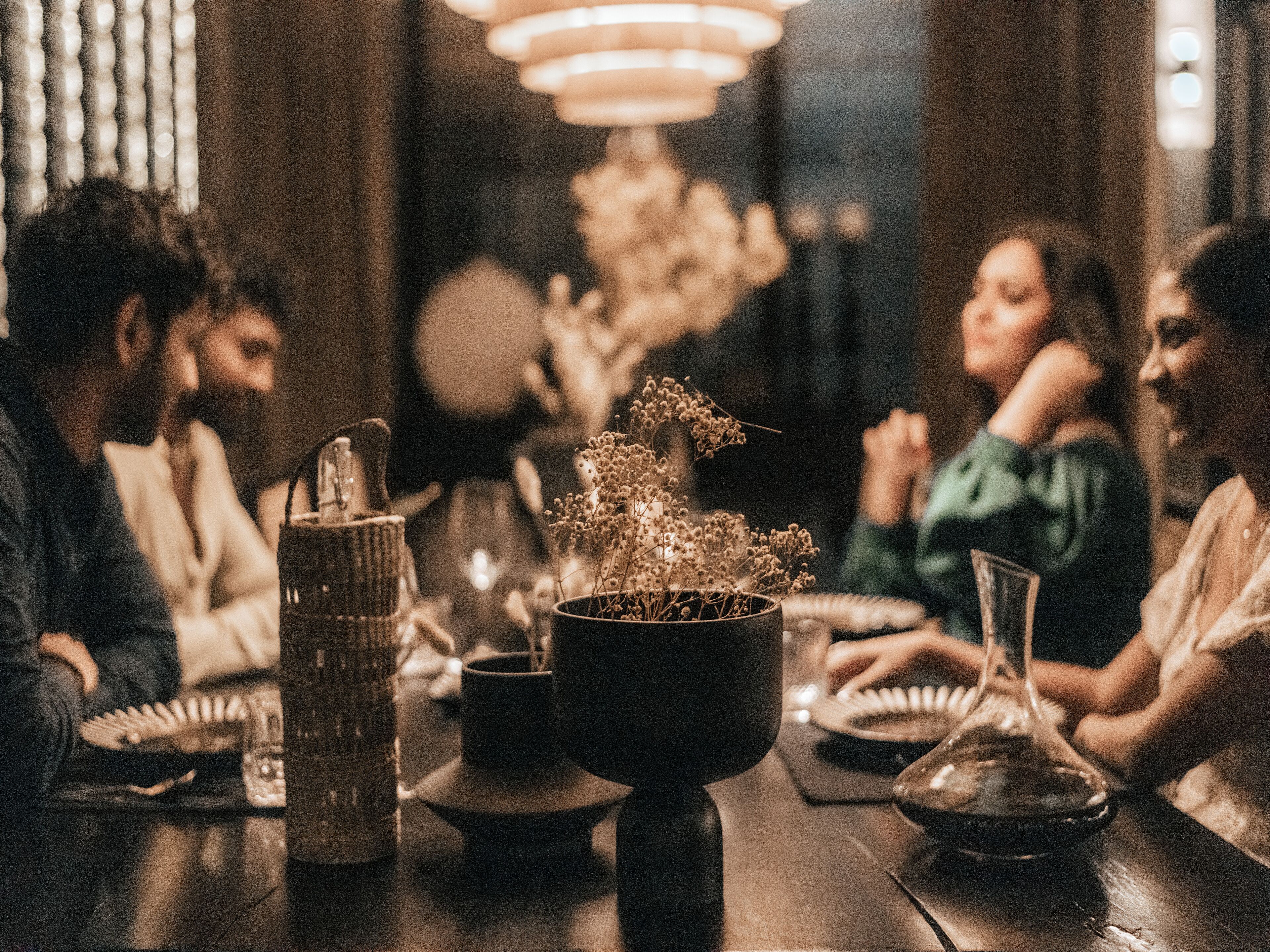 Un groupe de personnes est assis autour d’une table, devant une bougie, un vase rempli de fleurs séchées et un verre de vin.