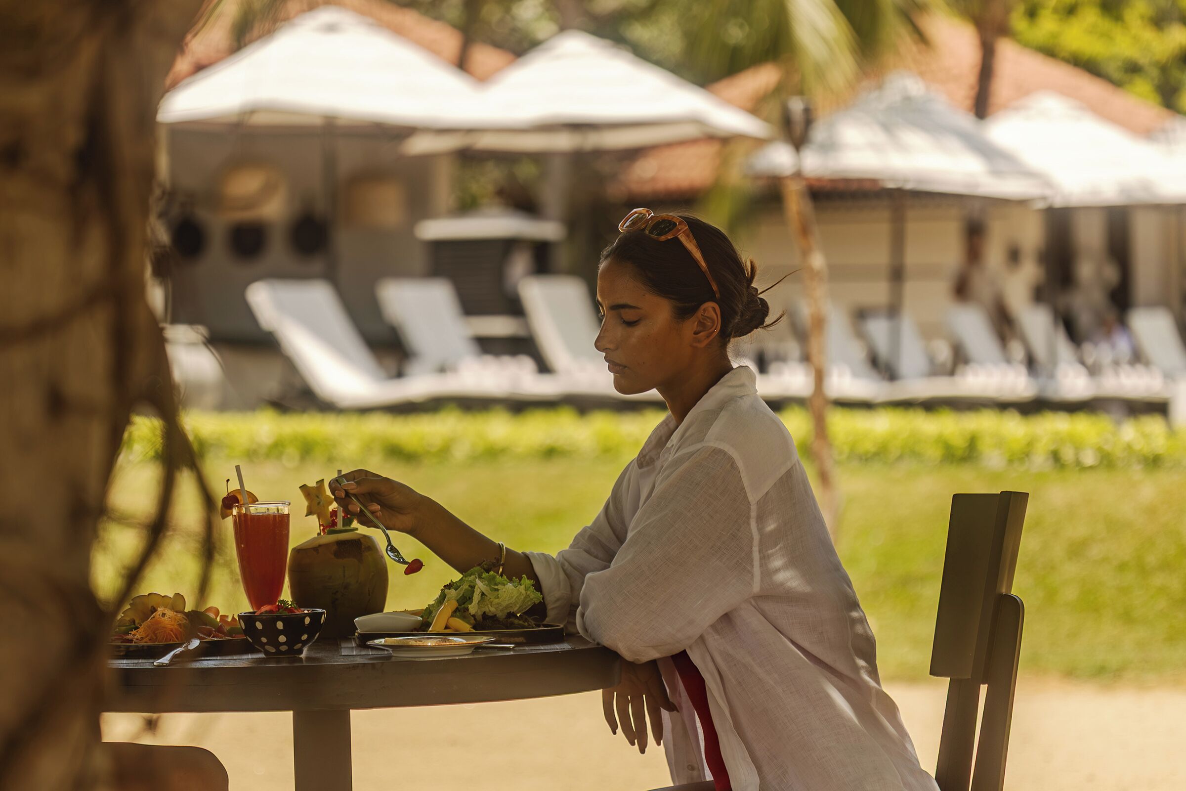 A person dining at an outdoor table with a drink and food, surrounded by lounge chairs and umbrellas.