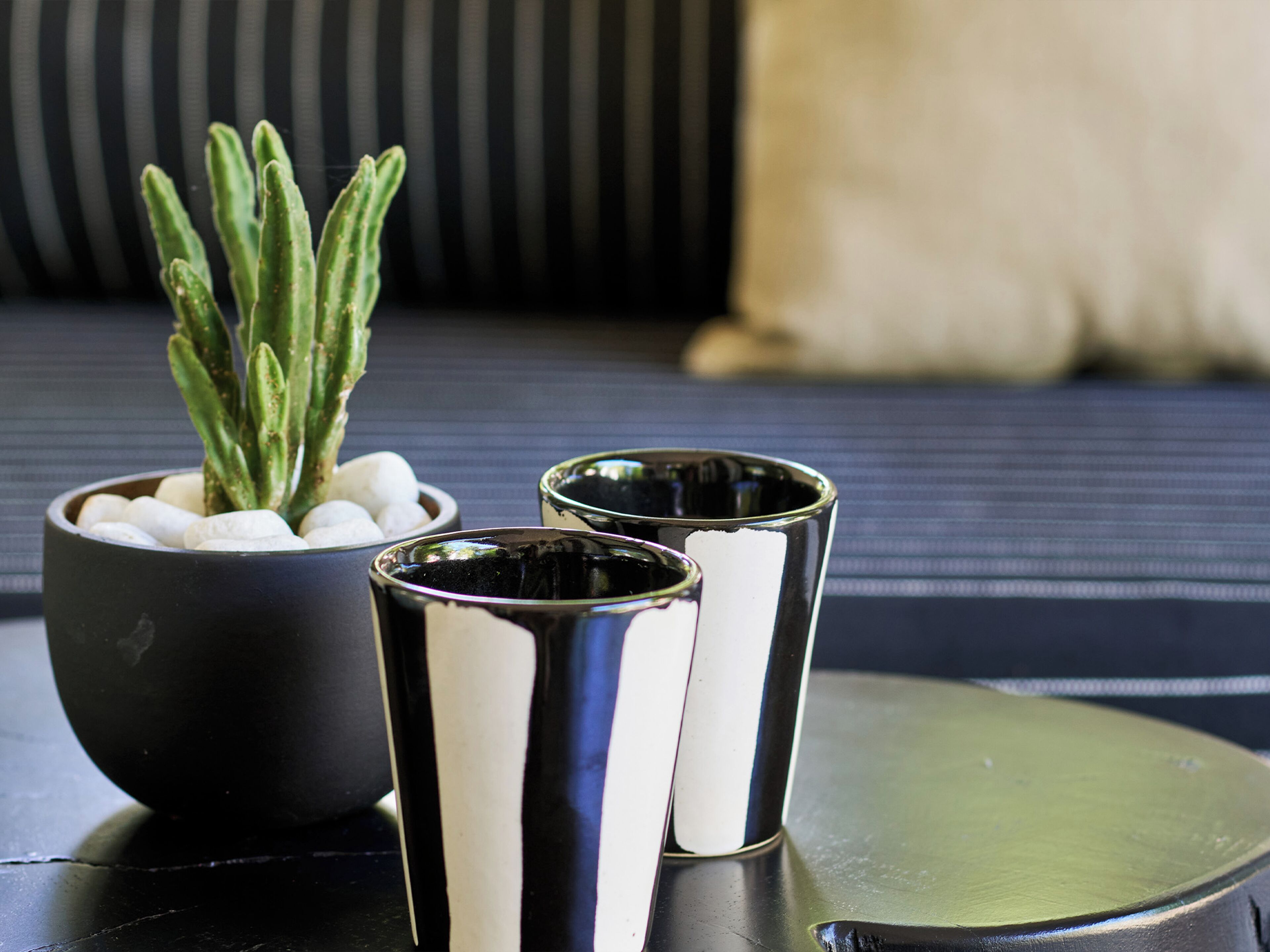 Een zwart-wit gestreepte bank met een kleine plant in pot en twee metalen bekers op een reflecterend oppervlak.