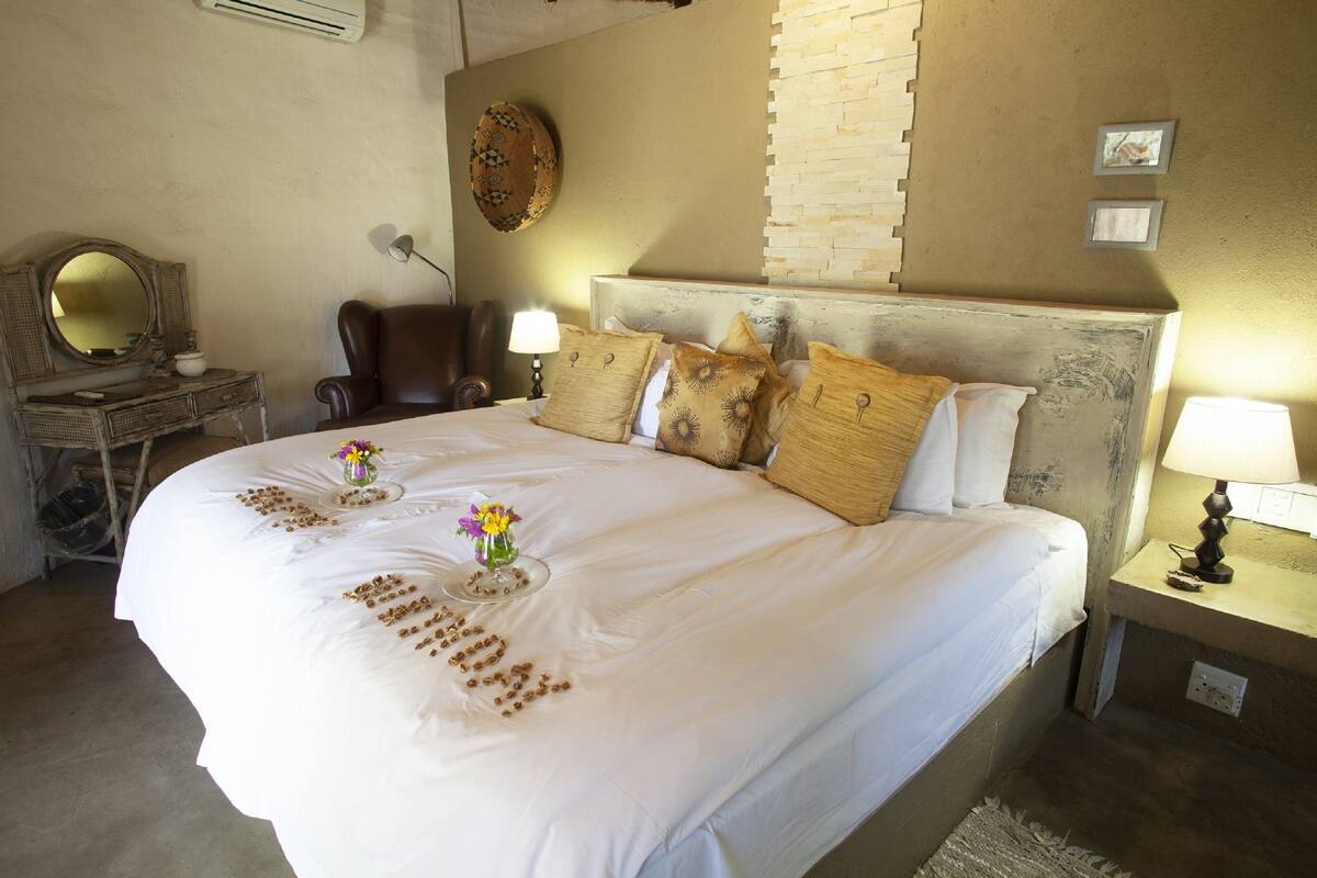 A neatly made bed with decorative pillows, a bedside table with a lamp, a chair, a mirror, and a wall-mounted decorative piece.
