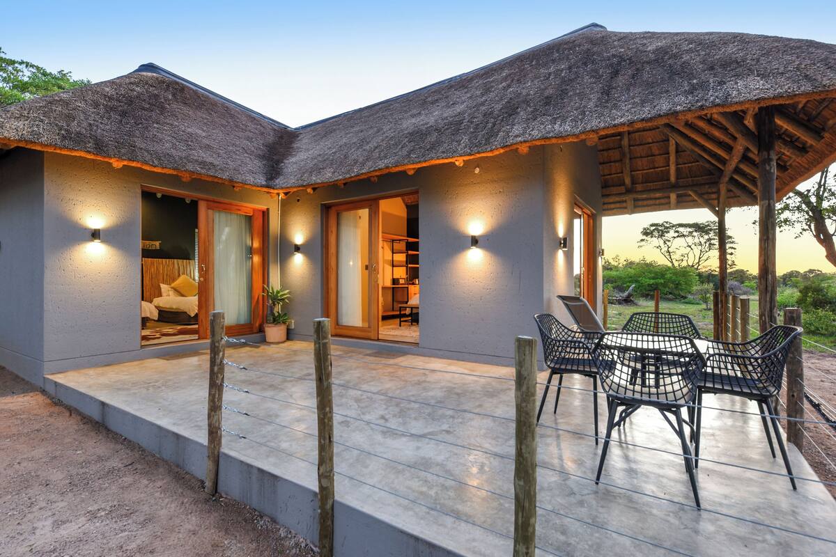 A modern building with a thatched roof, a covered patio with chairs, and a visible bedroom with a bed and a potted plant.
