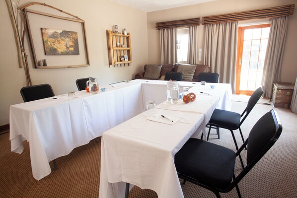 Un salón de conferencias con una mesa larga, sillas, un cuadro, una estantería y una ventana con cortinas.