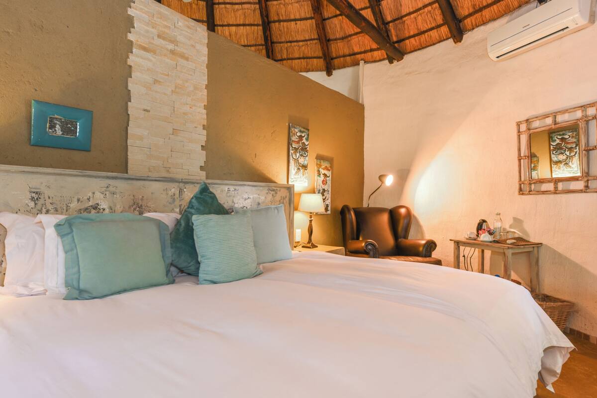 A bedroom with a bed, pillows, a chair, a lamp, a wall-mounted air conditioner, and a small table with items on it.