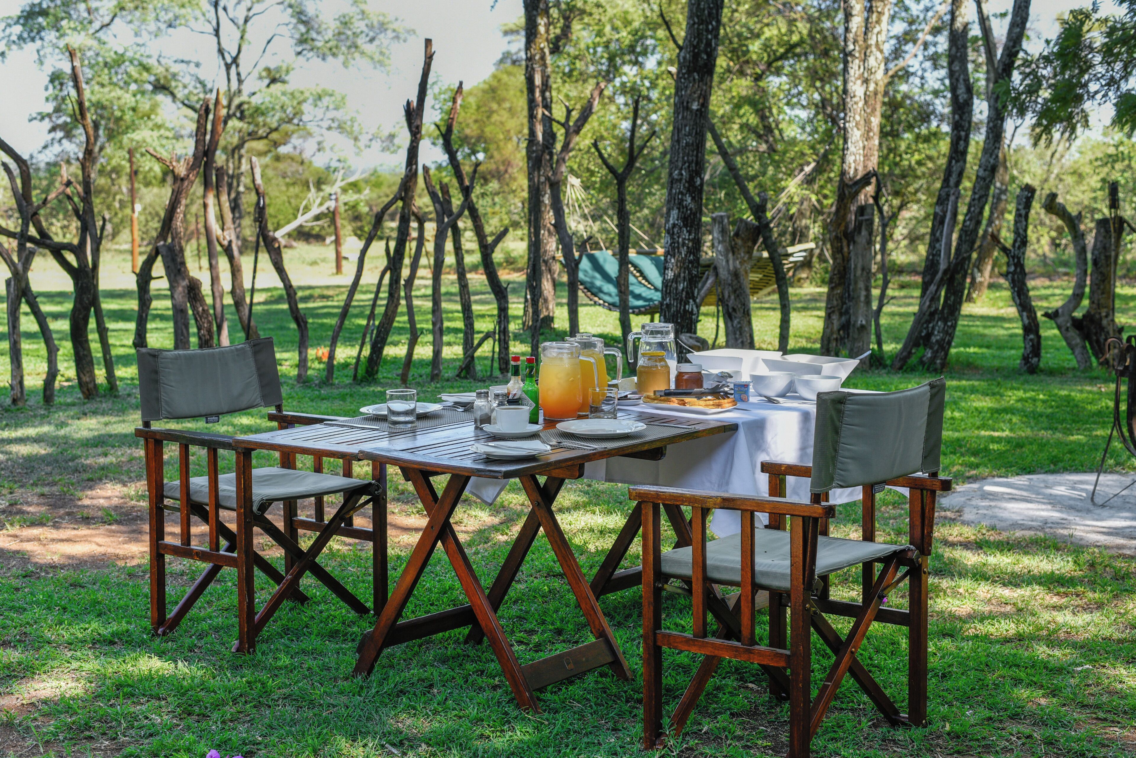 Un área de comedor al aire libre con una mesa preparada para dos, rodeada de sillas, y una zona verde con árboles al fondo.