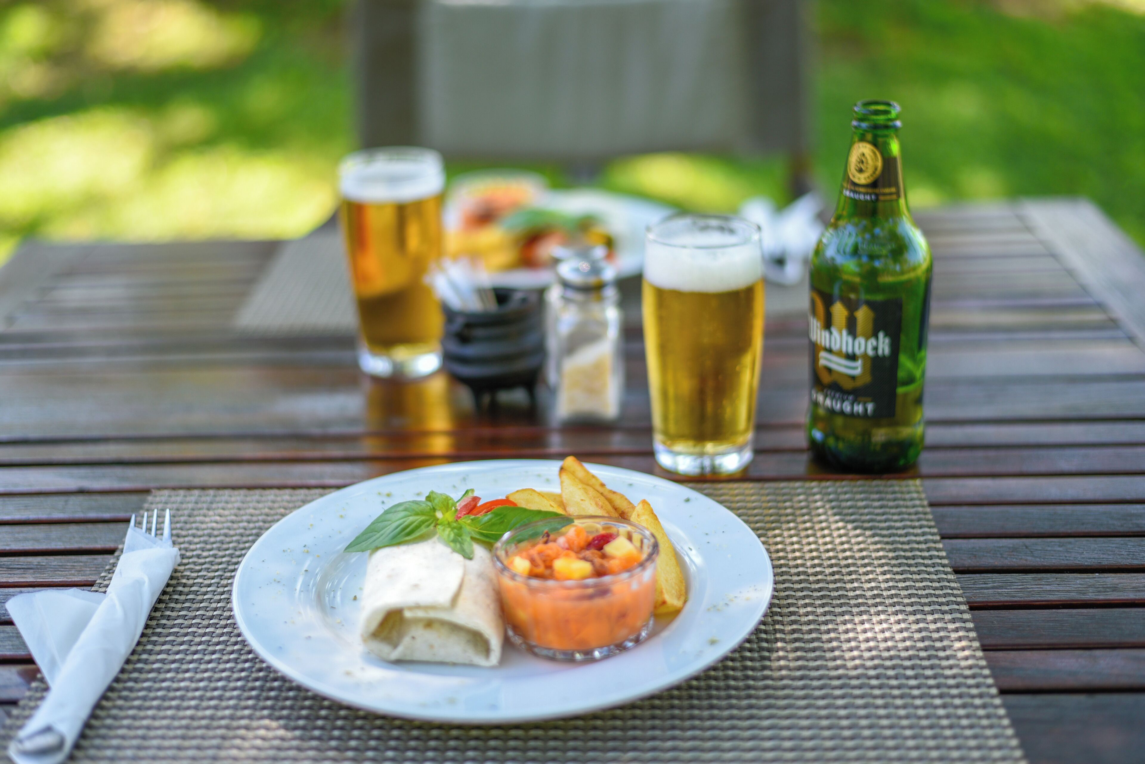 A dining table with a plate of food, a beer bottle, and glasses of beer.