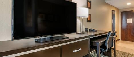 A hotel room with a large flat-screen TV, a desk with a chair, a lamp, and a door with a fire evacuation plan sign.