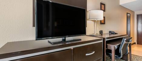 Ein Hotelzimmer mit Flachbildfernseher, Schreibtisch mit Lampe, Stuhl und Teppichboden.