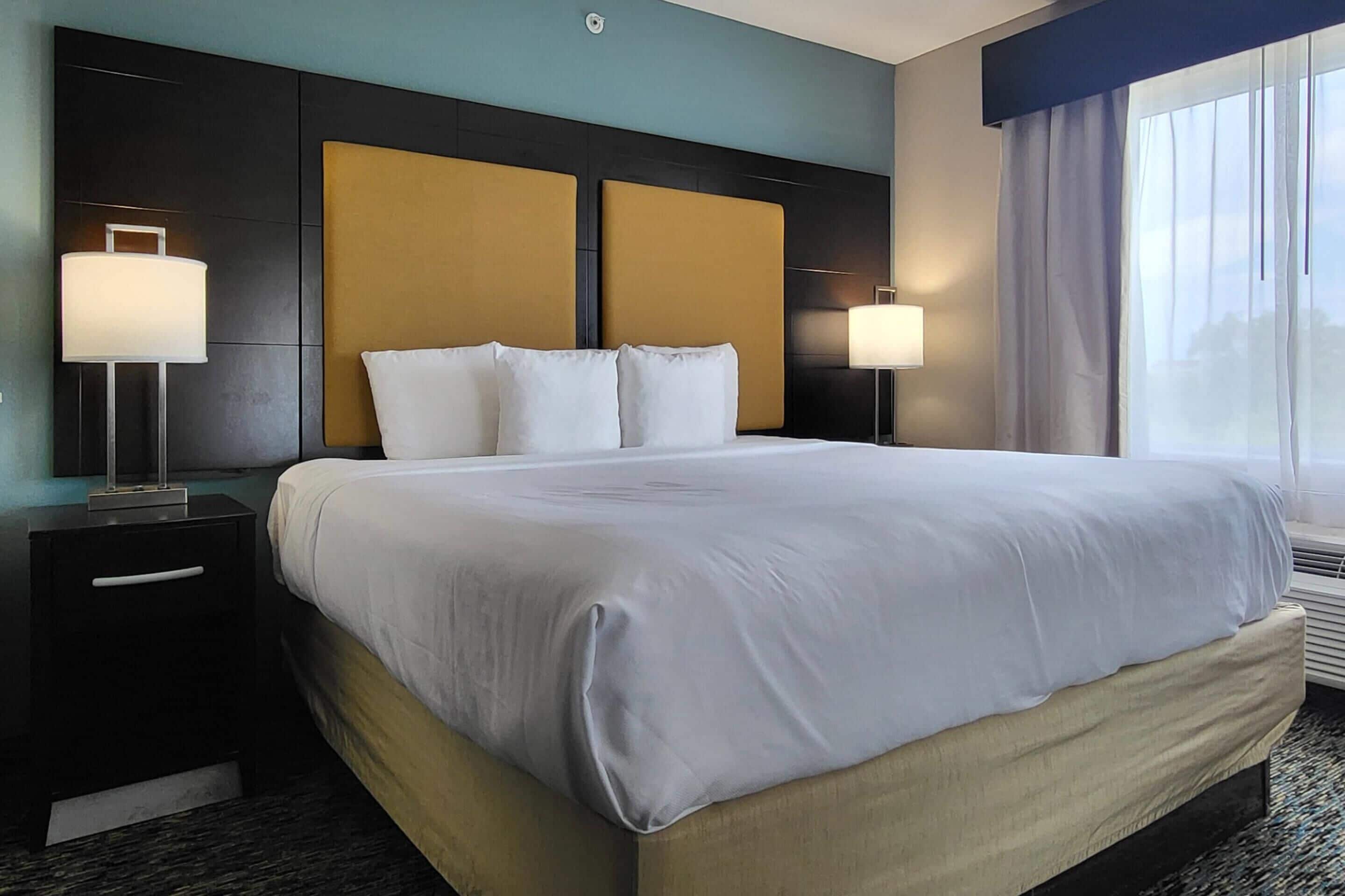 A hotel room with a large bed, two bedside tables with lamps, a headboard, and a window with curtains.