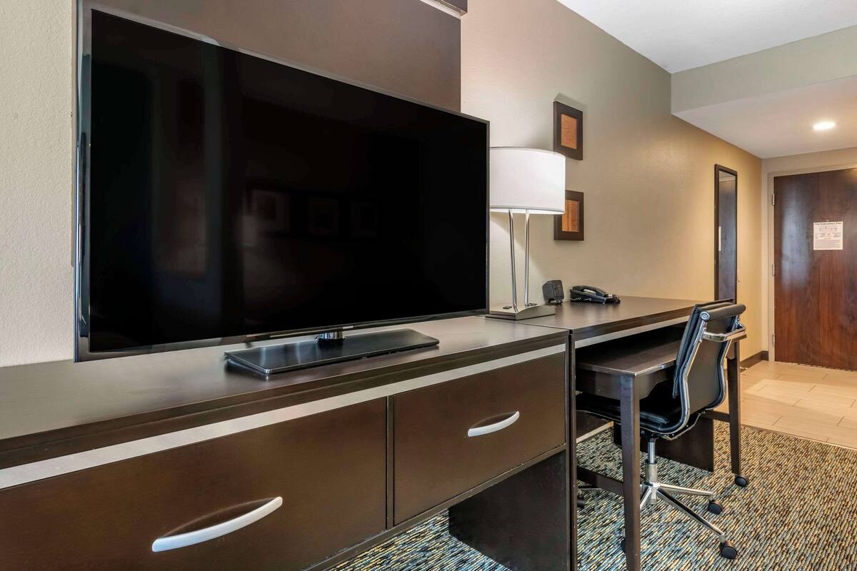 A hotel room with a large flat-screen TV mounted on a dark wooden dresser, a desk with a chair, a lamp, and a door with a sign.