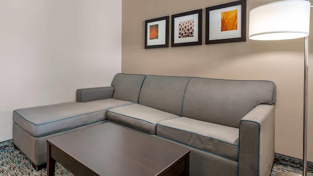 A hotel room with a gray sectional sofa, a small wooden coffee table, and three framed artworks on the wall.