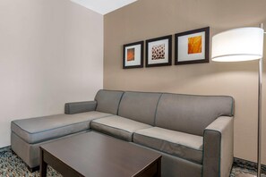Habitación de hotel con un sofá seccional gris, una pequeña mesa de centro de madera y tres obras de arte enmarcadas en la pared.