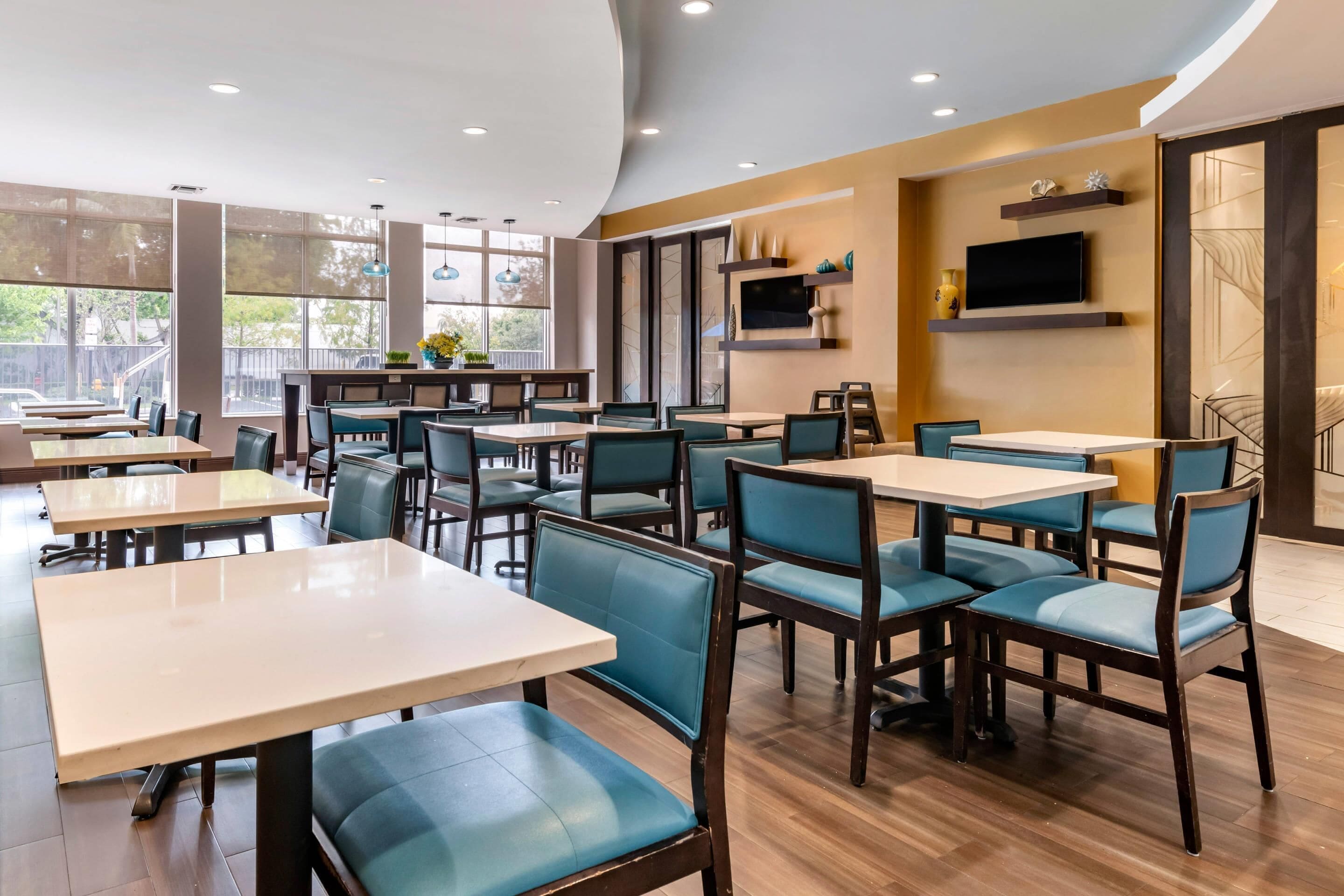 Un interior de restaurante moderno con varias mesas y sillas, ventanas amplias y un televisor de pantalla plana en la pared.