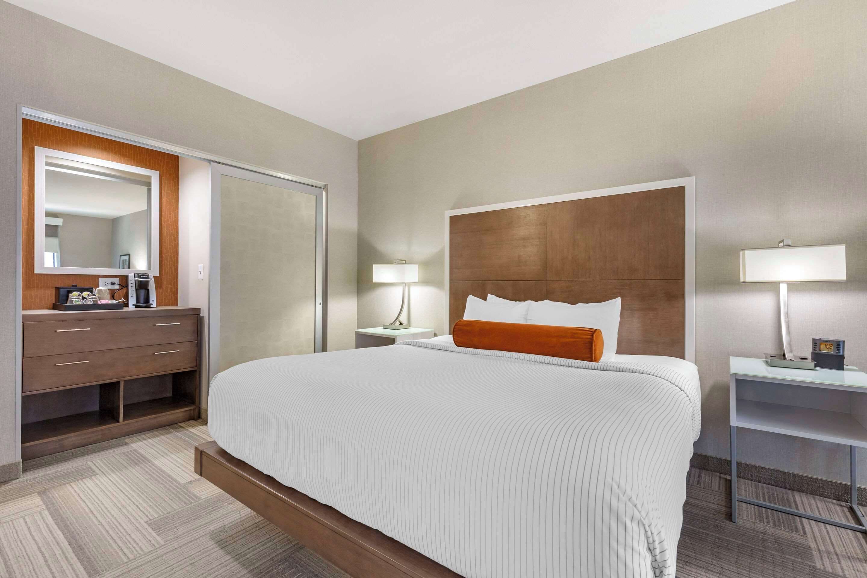 A hotel room with a large bed, a wooden headboard, a dresser, a mirror, two bedside tables with lamps, and a carpeted floor.