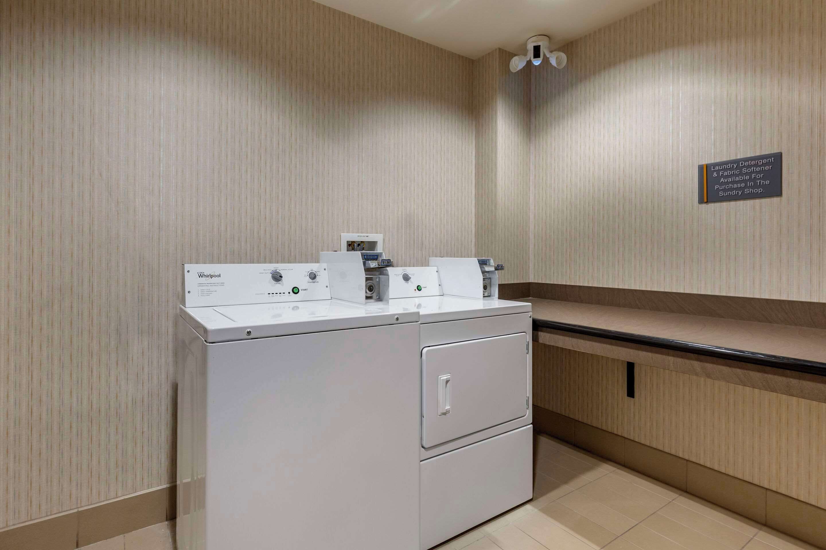Een wasruimte met een Whirlpool wasmachine en droger, een bordje dat aangeeft waar wasmiddel en wasverzachter verkrijgbaar zijn, en een aanrecht met een wasbak.