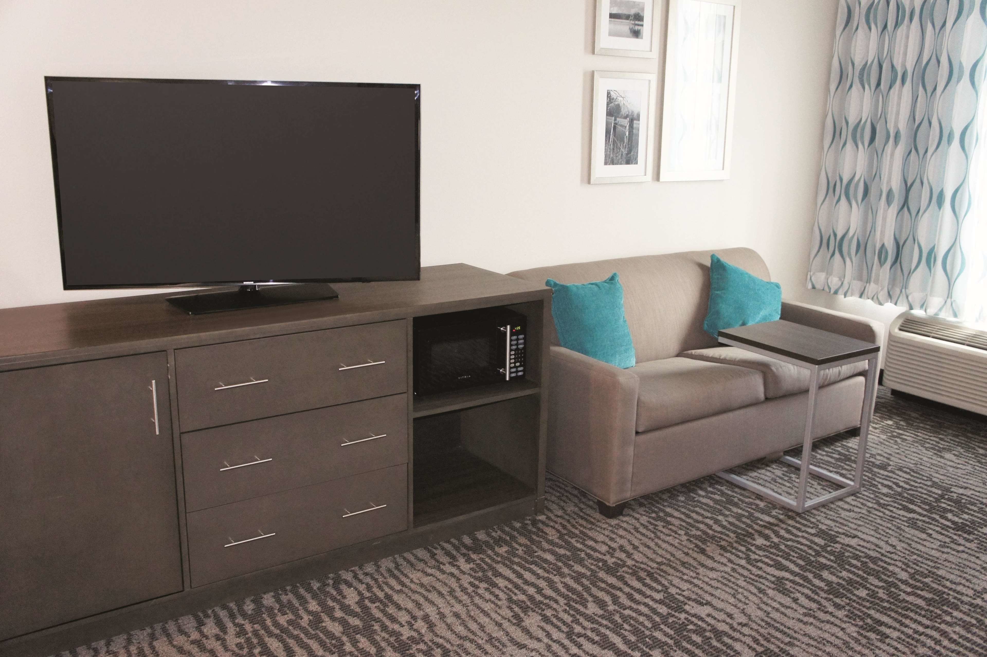Habitación de hotel con televisor de pantalla plana sobre un mueble de madera, sofá gris con cojines verde azulado, mesita auxiliar pequeña y alfombra estampada.