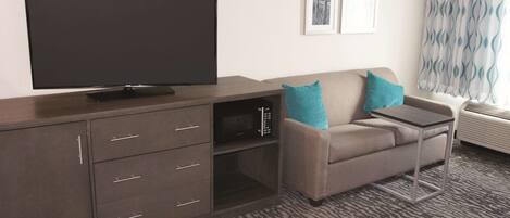 Habitación de hotel con televisor de pantalla plana sobre un mueble de madera, sofá gris con cojines verde azulado, mesita auxiliar pequeña y alfombra estampada.