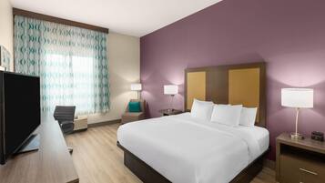A hotel room with a large bed, a desk with a flat-screen TV, two bedside lamps, a chair, and a window with patterned curtains.