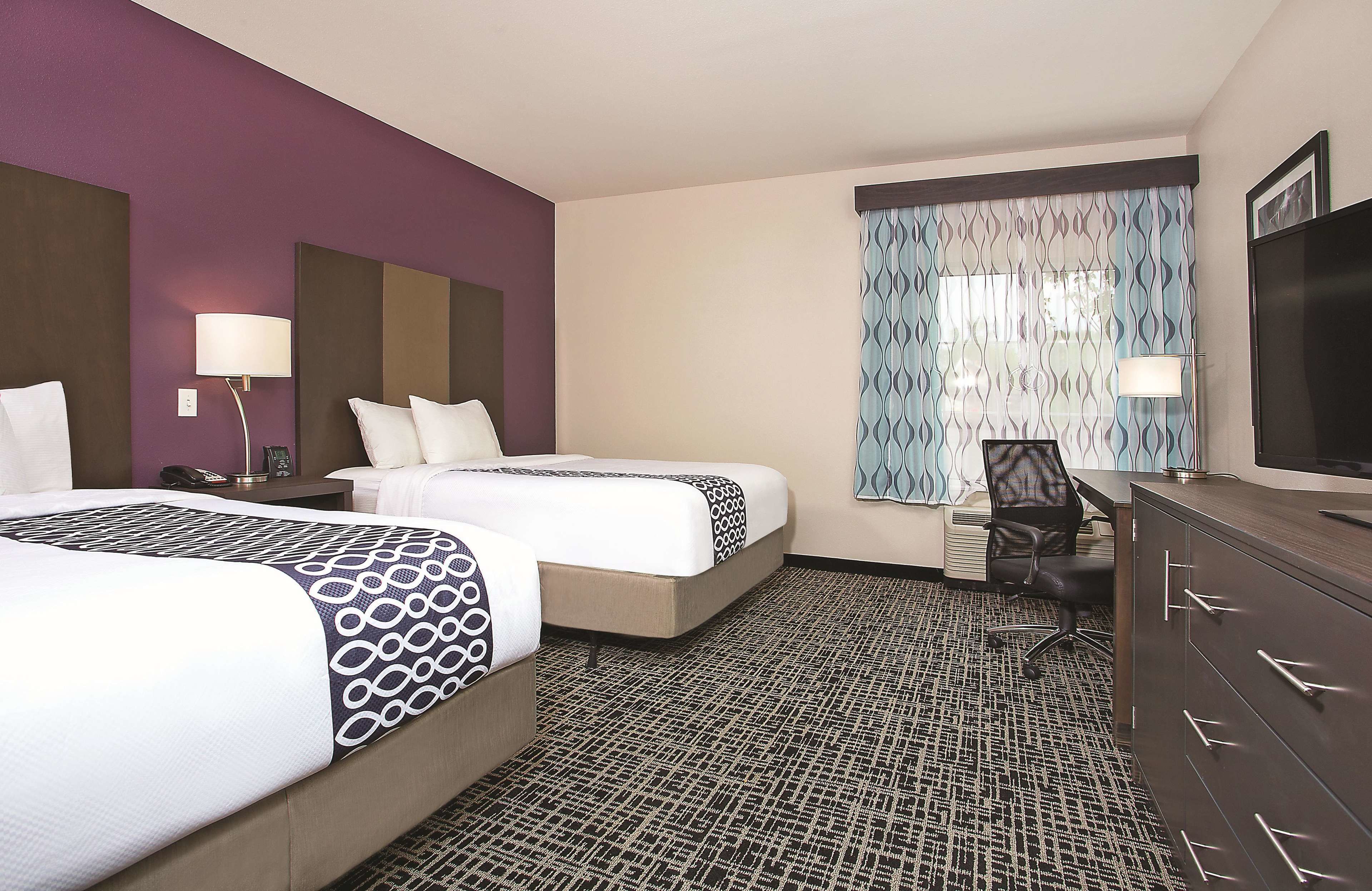 A hotel room with two beds, a desk, a chair, a TV, and a patterned carpet.