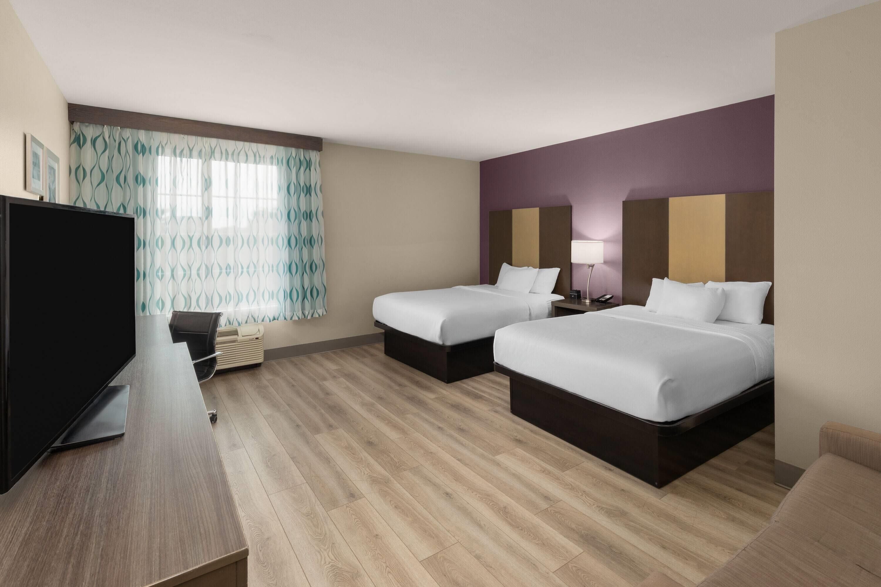 Ein Hotelzimmer mit zwei Betten, einem Fernseher, einem Schreibtisch und einem Fenster mit gemusterten Vorhängen.