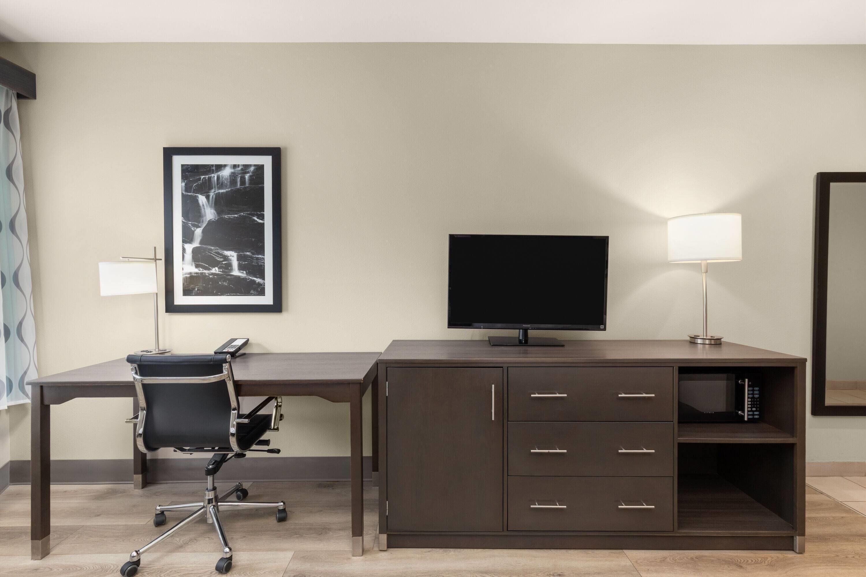 A hotel room with a dark wooden desk, a black office chair, a flat-screen TV, a lamp, a framed picture on the wall, and a mirror.
